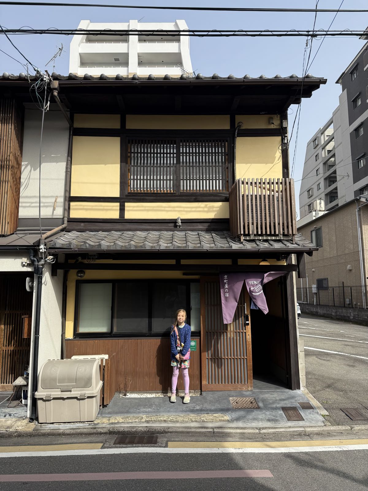 A child stands in front of a traditional Japanese house. The house is brown and yellow, and has two storeys.