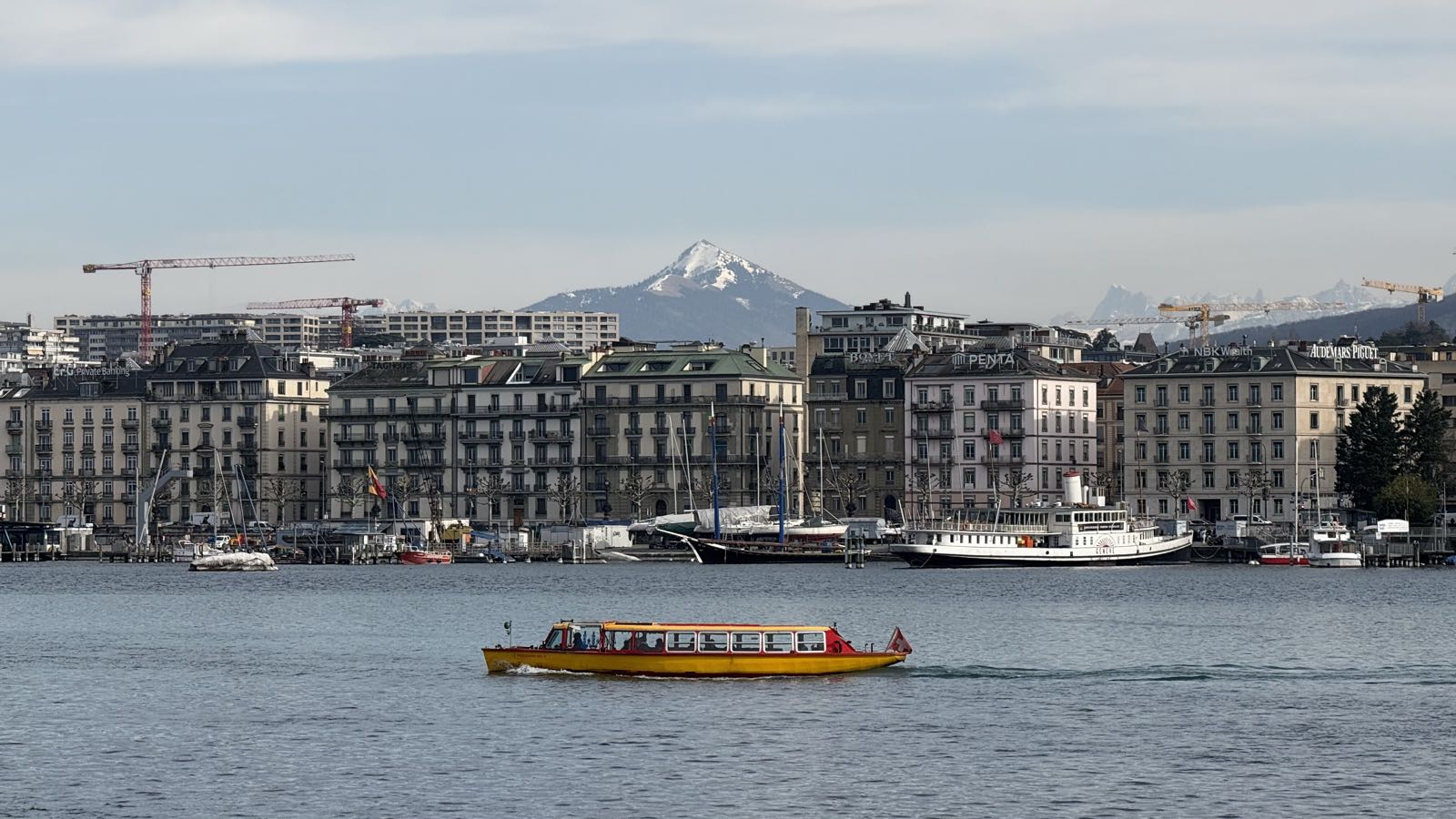 Between a lake and a sunny sky, a portion of Geneva’s skyline is visible. Mont-Blanc stands behind the city, and in the lake, a yellow tourist boat goes by.