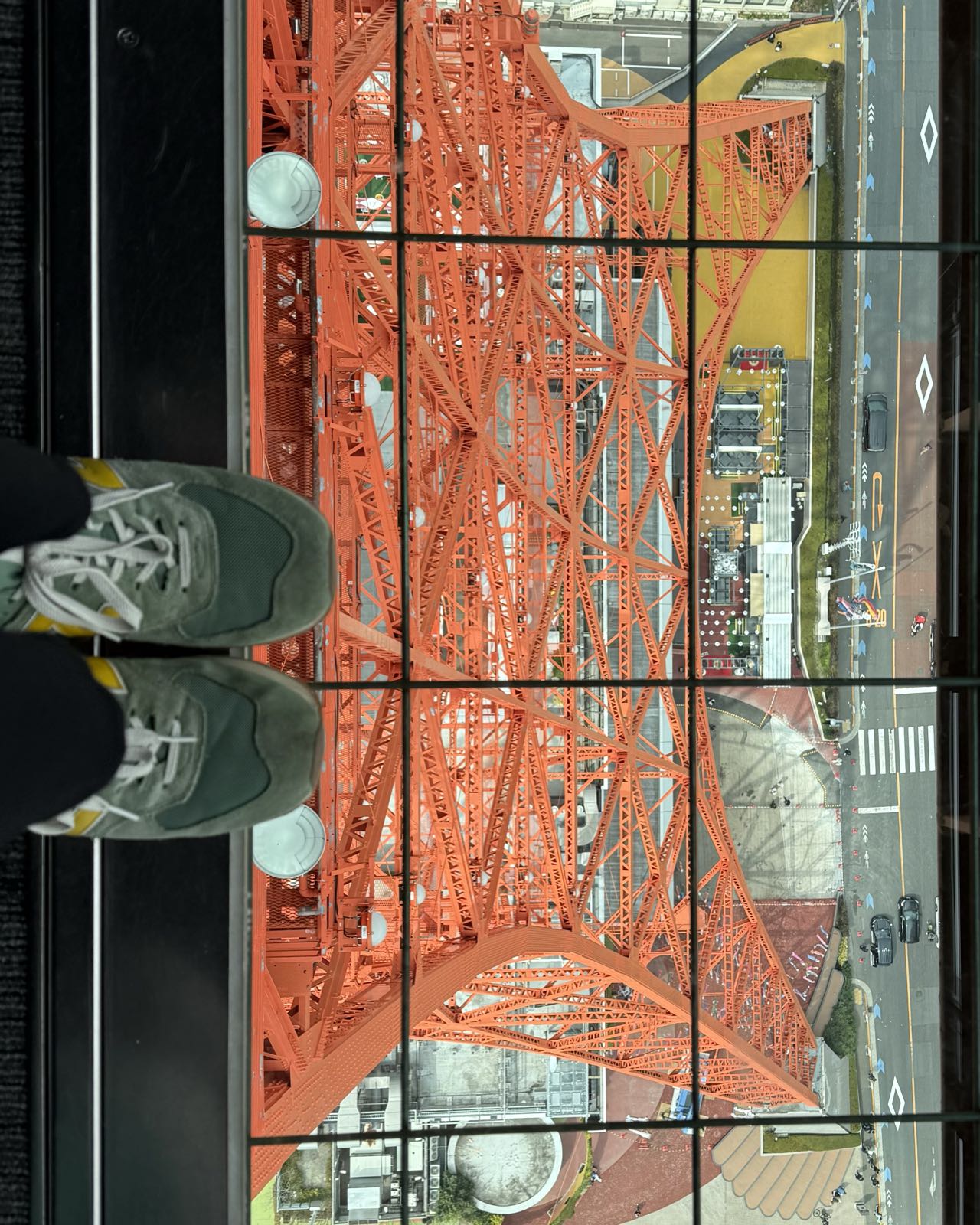 Two feet wearing green sneakers stick into a window in the floor of a room. Through the window, the orange lower section of Tokyo Tower is visible, as is the street below.