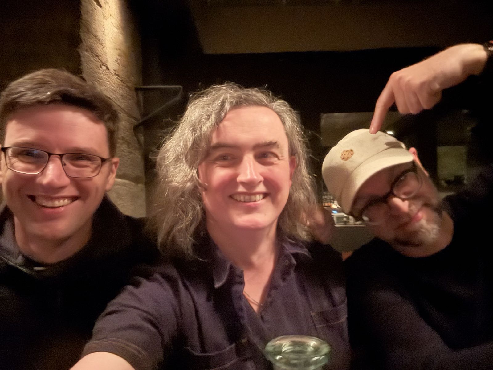 A somewhat blurry photo of three people sitting next to each other at a bar. The smiling man on the left is clean-shaven. The woman in the middle is the one taking the photo, with the selfie camera on her phone. She has shoulder-length wavy hair, and is also smiling. The man on the right is wearing a baseball cap, and is pointing at the woman in a somewhat strange manner.