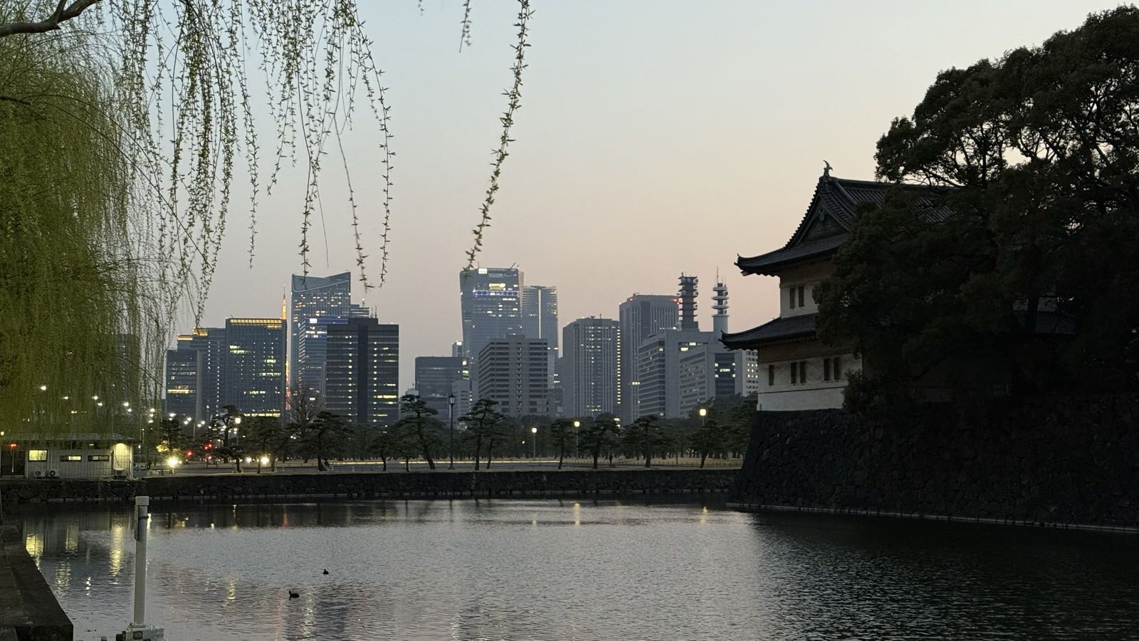 A city skyline at dusk. In front of the skyline is a body of water. To the right of the water is a white building with a black roof, done in traditional Japanese architecture.