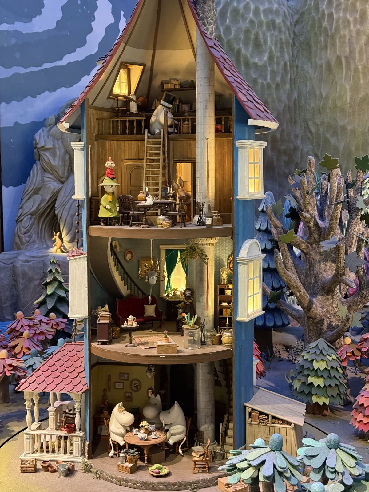 Within a model of a wooded valley, a cut-away model of a circular, four storey house stands. Within the house are various white blobular creatures—Moomins—engaged in various activities.