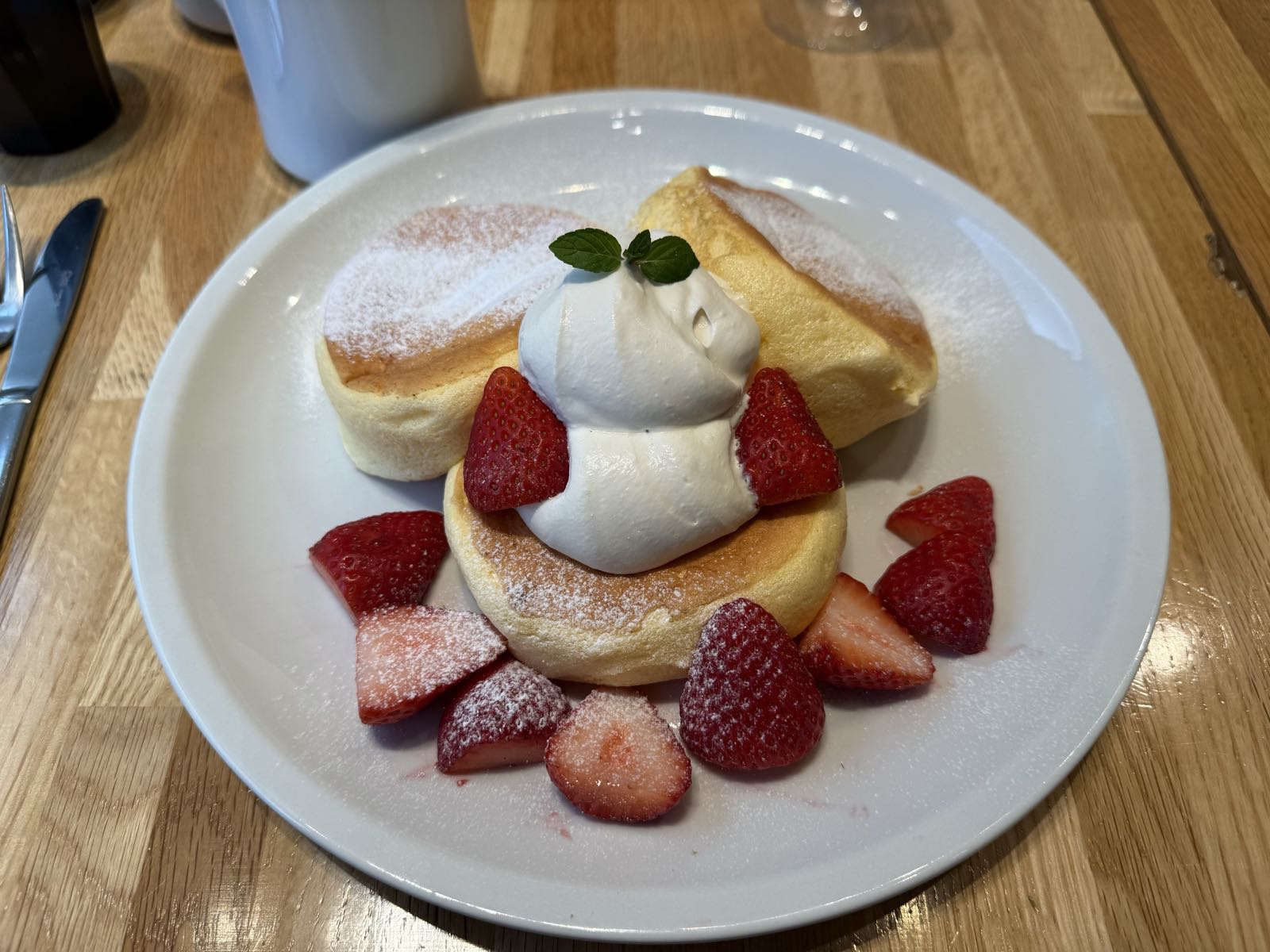 Three kiseki (souflée) pancakes sit on a plate. a large dollop of cream sits on the one in the center. All three have powdered sugar on them, and the plate has a dozen strawberries artfully arrayed around and on the pancakes.