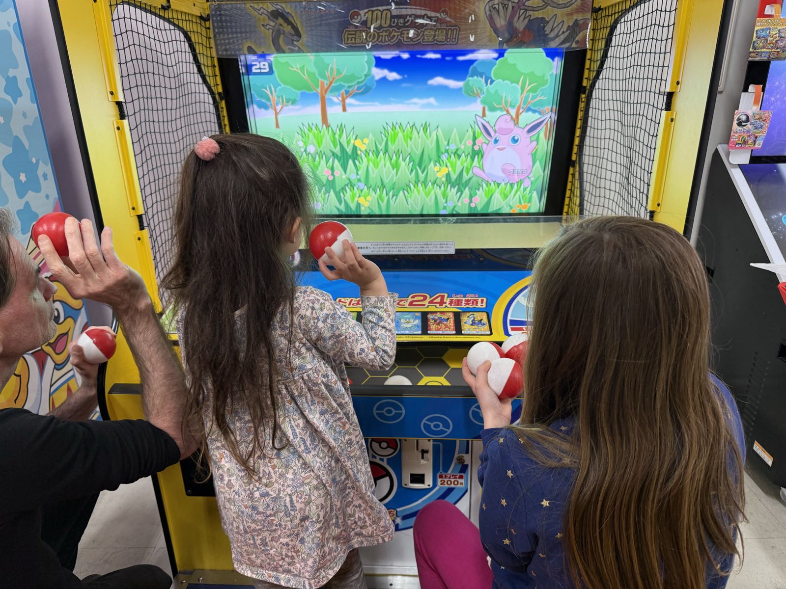 Two children and one adult are holding pokéballs and watching a video screen depicting a pink Pokémon standing in a grassy field.