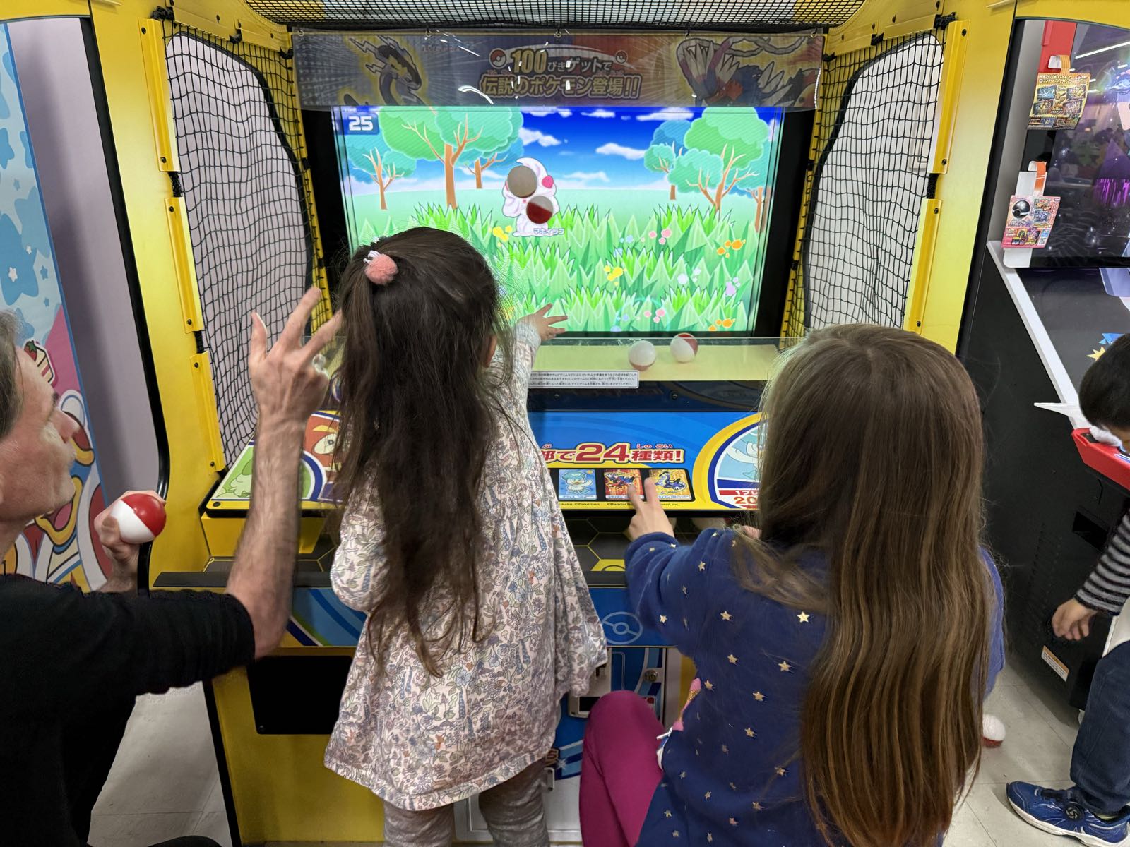 Two children and one adult are throwing pokéballs at a video screen depicting a grassy field.