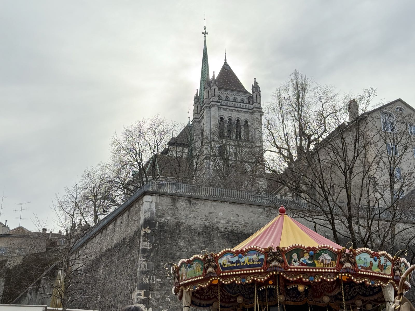 An old stone cathedral on a hill is above and behind a stone house and wall. In front of the wall, downhill from both the cathedral and the house, is a carousel.