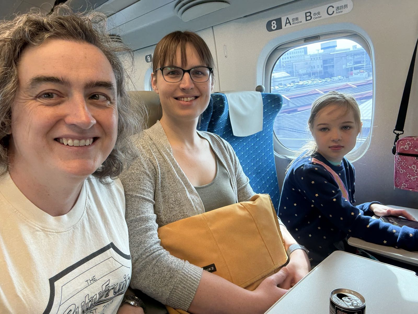 Two women and a girl sit next to each other on a train. The two women are smiling at the camera. The child is giving the camera some RBF. The woman in the center has a large yellow bag.