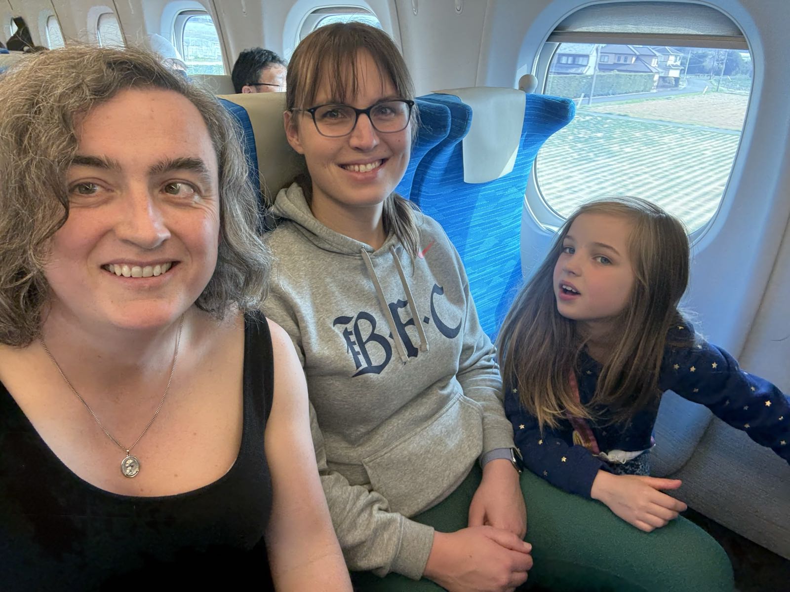 Two women and a girl sit next to each other on a train. The two women are smiling at the camera. The child is giving the camera some side-eye.