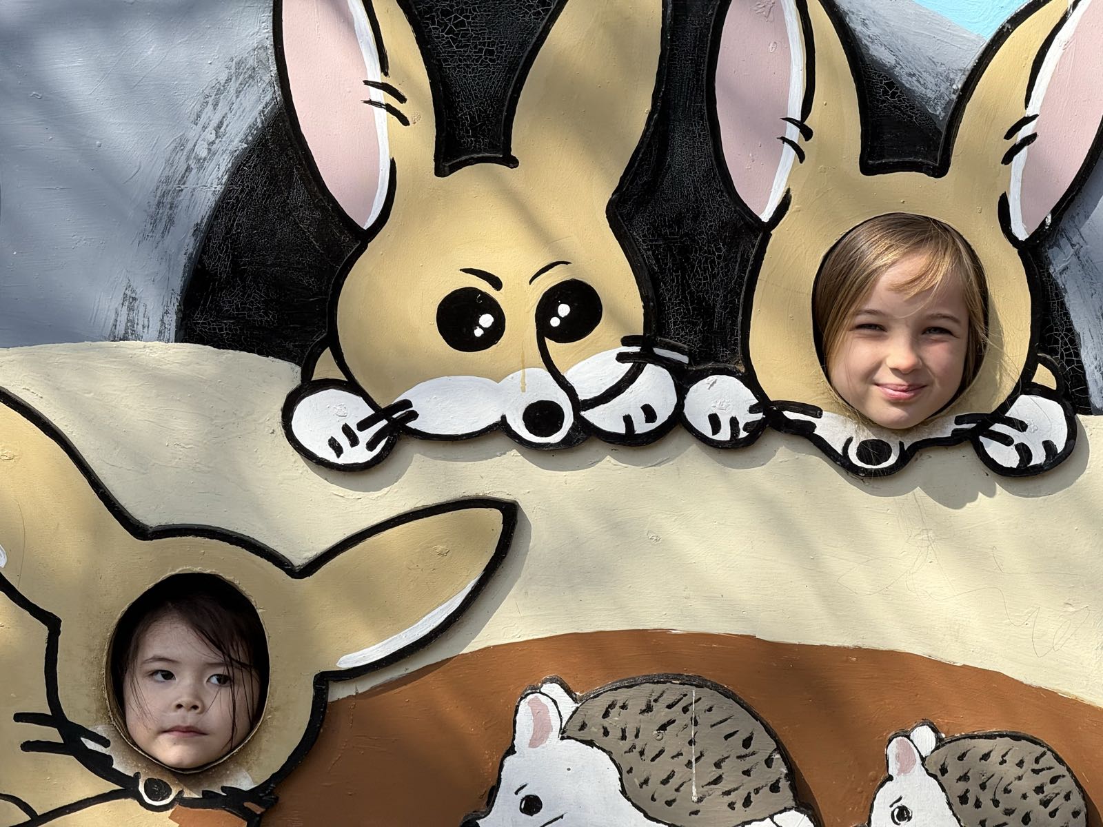 Two children pose for a photo at a sign for Inokashira Park Zoo. The sign has holes in it at various animal faces, and the children’s faces are visible in the holes, so the children appear to be various zoo animals.
