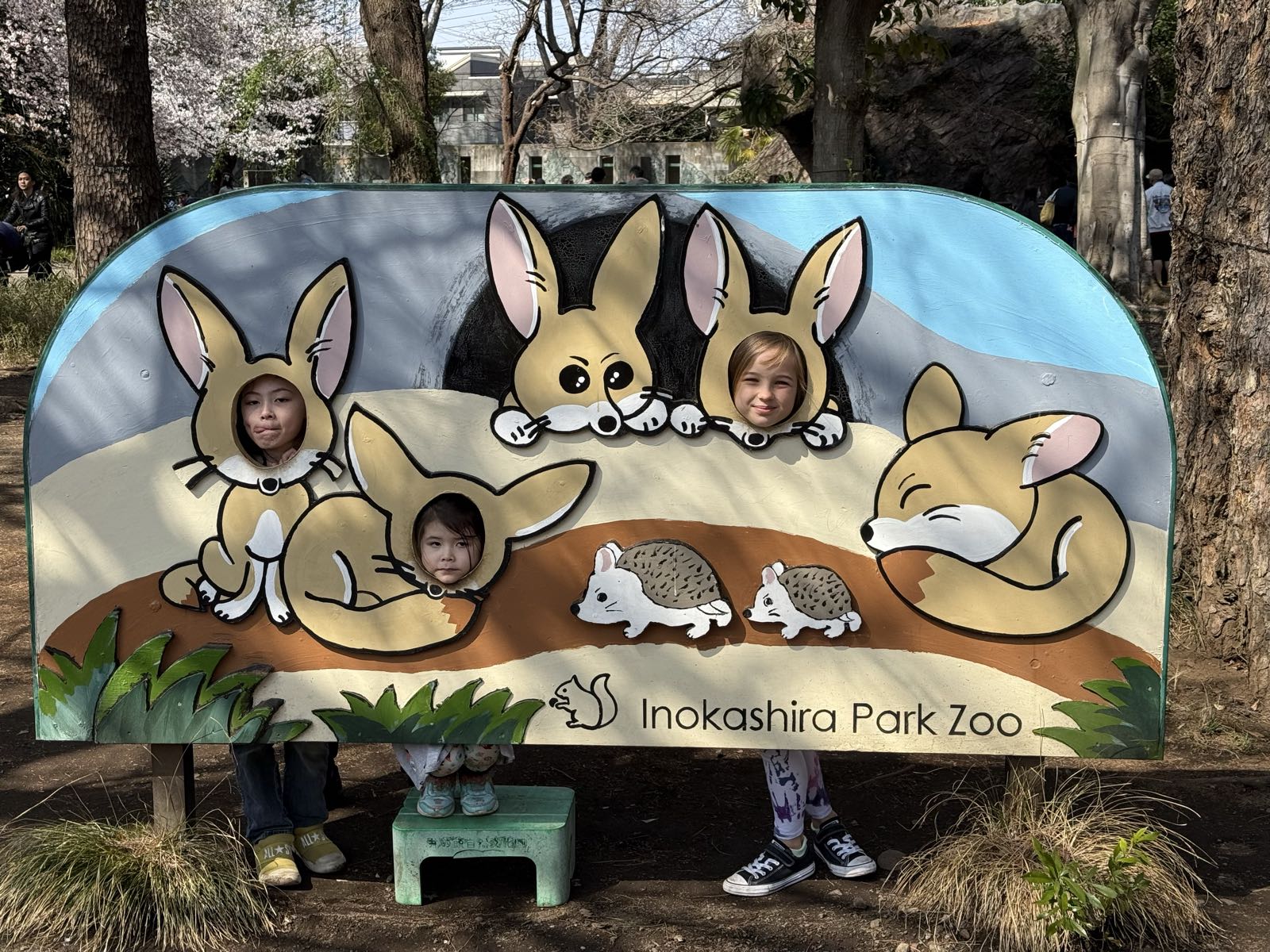 Three children pose for a photo at a sign for Inokashira Park Zoo. The sign has holes in it at various animal faces, and the children’s faces are visible in the holes, so the children appear to be various zoo animals.