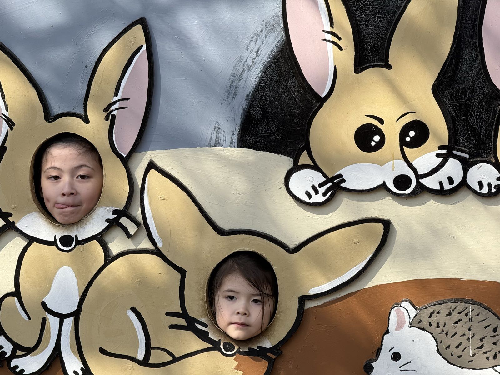 Two children pose for a photo at a sign for Inokashira Park Zoo. The sign has holes in it at various animal faces, and the children’s faces are visible in the holes, so the children appear to be various zoo animals.
