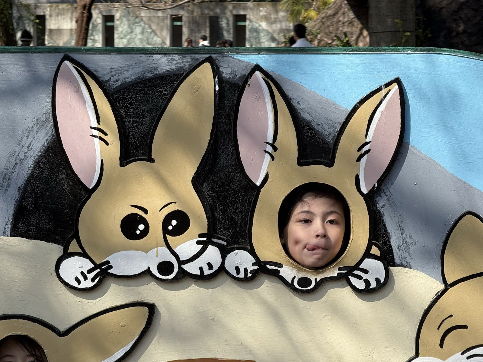 A child poses for a photo at a sign for Inokashira Park Zoo. The sign has holes in it at various animal faces, and the child’s face is visible in one of the holes, so the child appears to be a zoo animal.