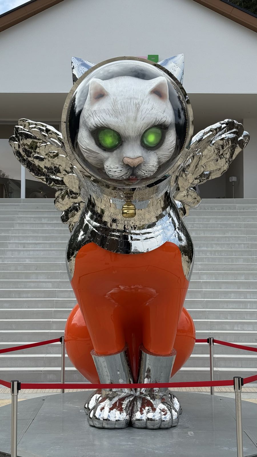 A large statue of a white, winged cat wearing an orange spacesuit. The cat has glowing green orbs for eyes.