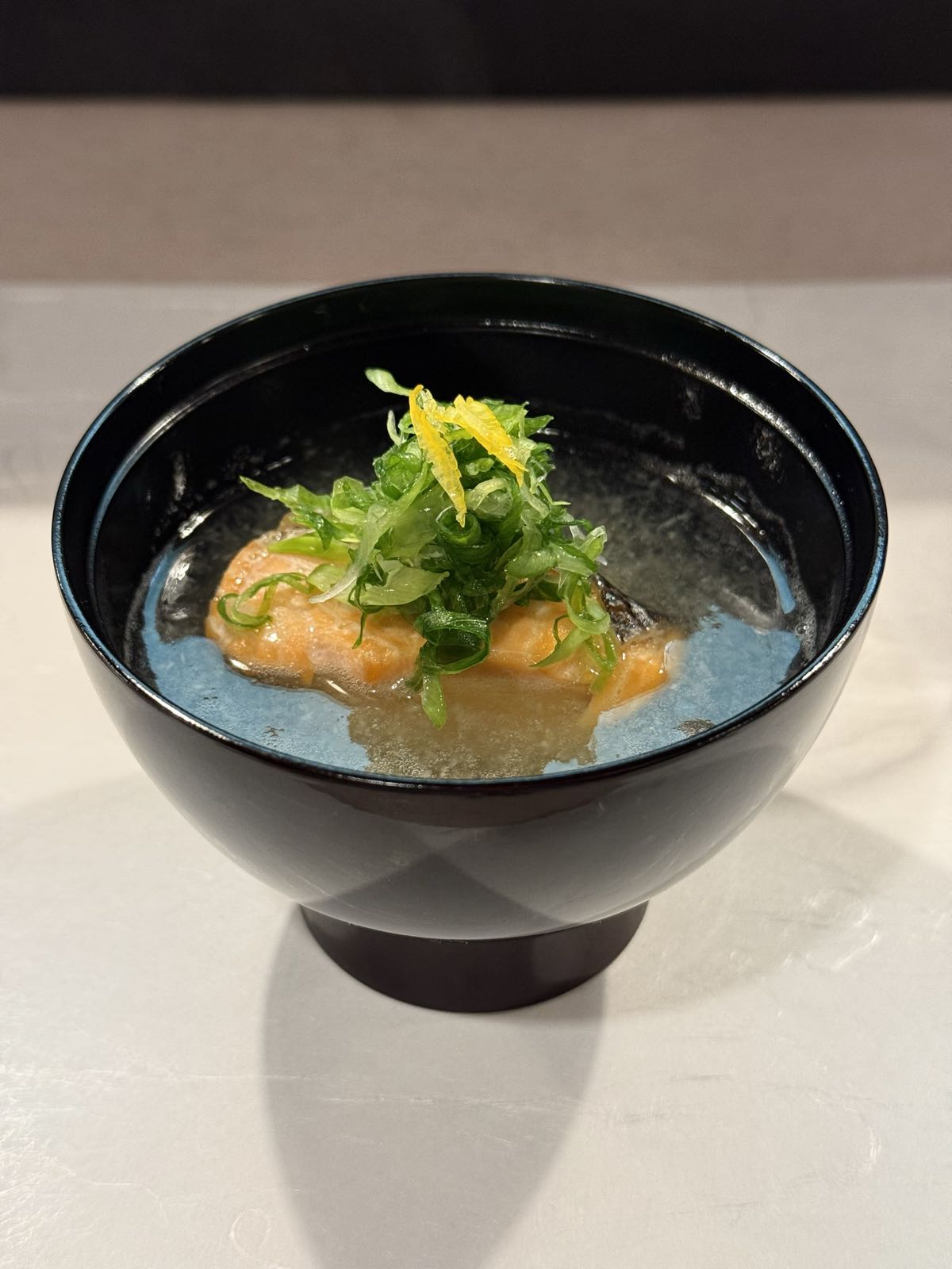 A piece of salmon sits in a bowl of miso soup. There is a tidy pile of green onion slices atop it.