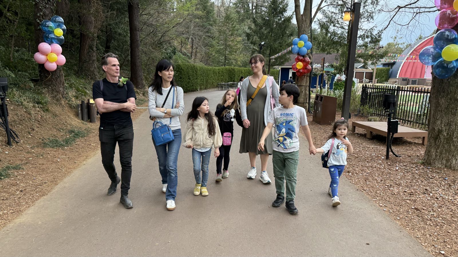Three adults and four children walk along a paved path among trees decorated with colorful balloons. Some are holding hands. Everybody looks a bit tired.