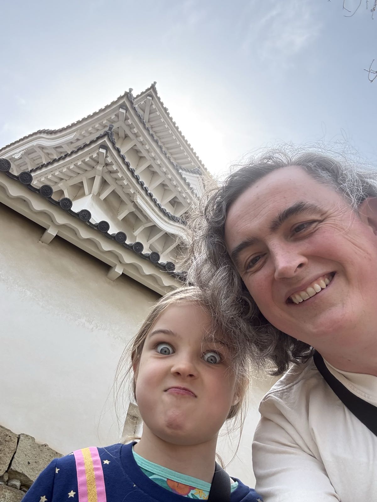 Below a white Japanese castle, a child and a woman pose for a selfie. The woman is smiling, while the child is making a very silly face.
