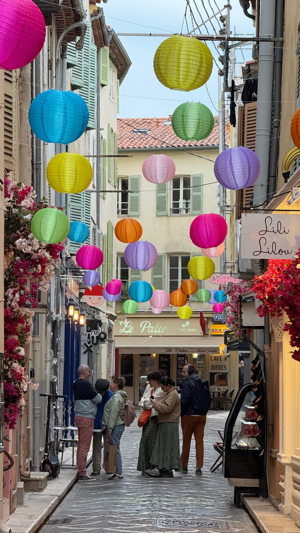 A cobblestone street lined with restaurants and shops stretches away from the camera. Suspended above the street are many colorful paper lanterns. A group of people stand on the street in the center of the frame.