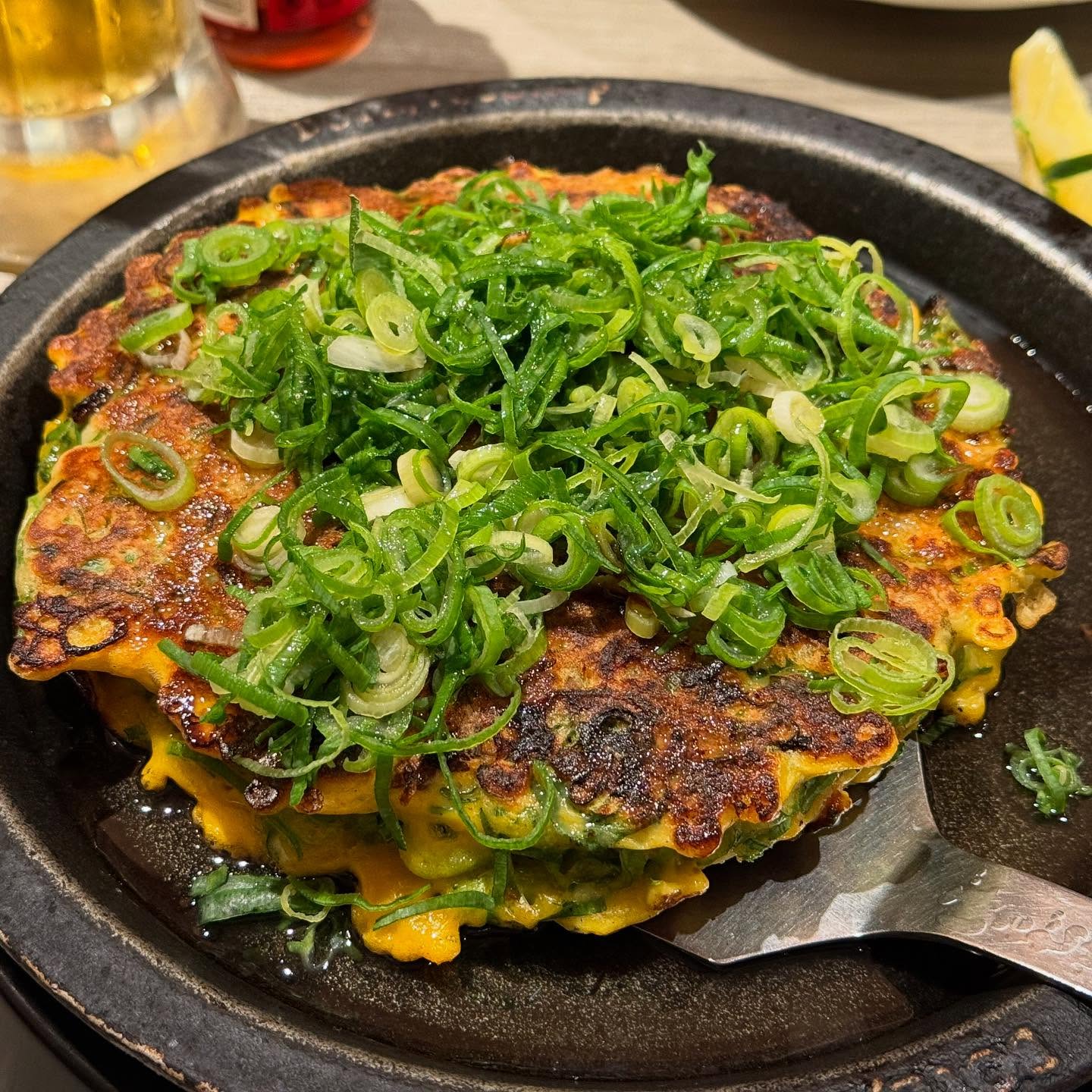 A cast-iron pan has okonomiyaki in it. The pancake is covered in sliced green onion. A metal spatula is tucked underneath it.