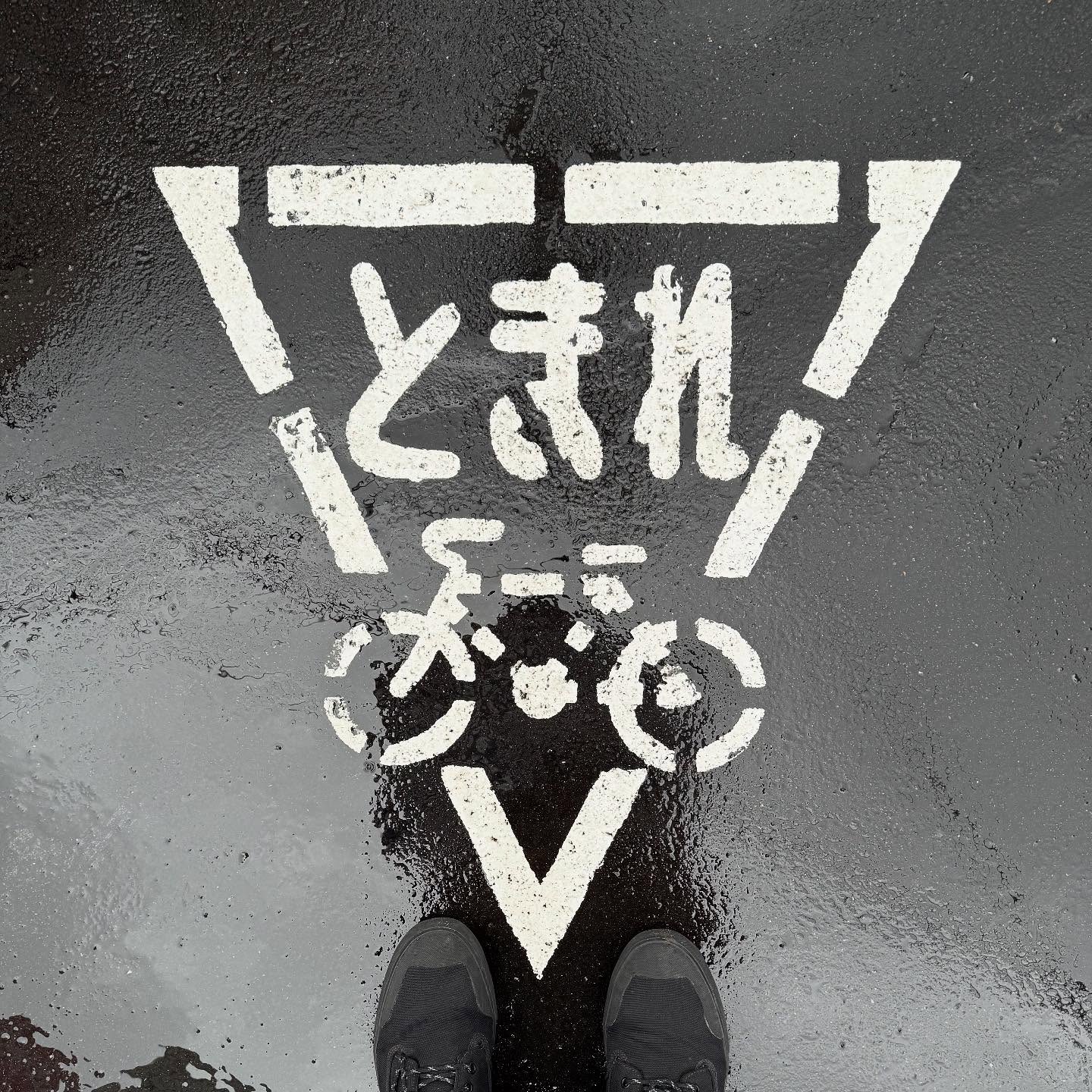 A white, triangular road marking depicts a bicycle and reads 「とまれ」. Two feet in black boots are visible at the bottom of the frame.