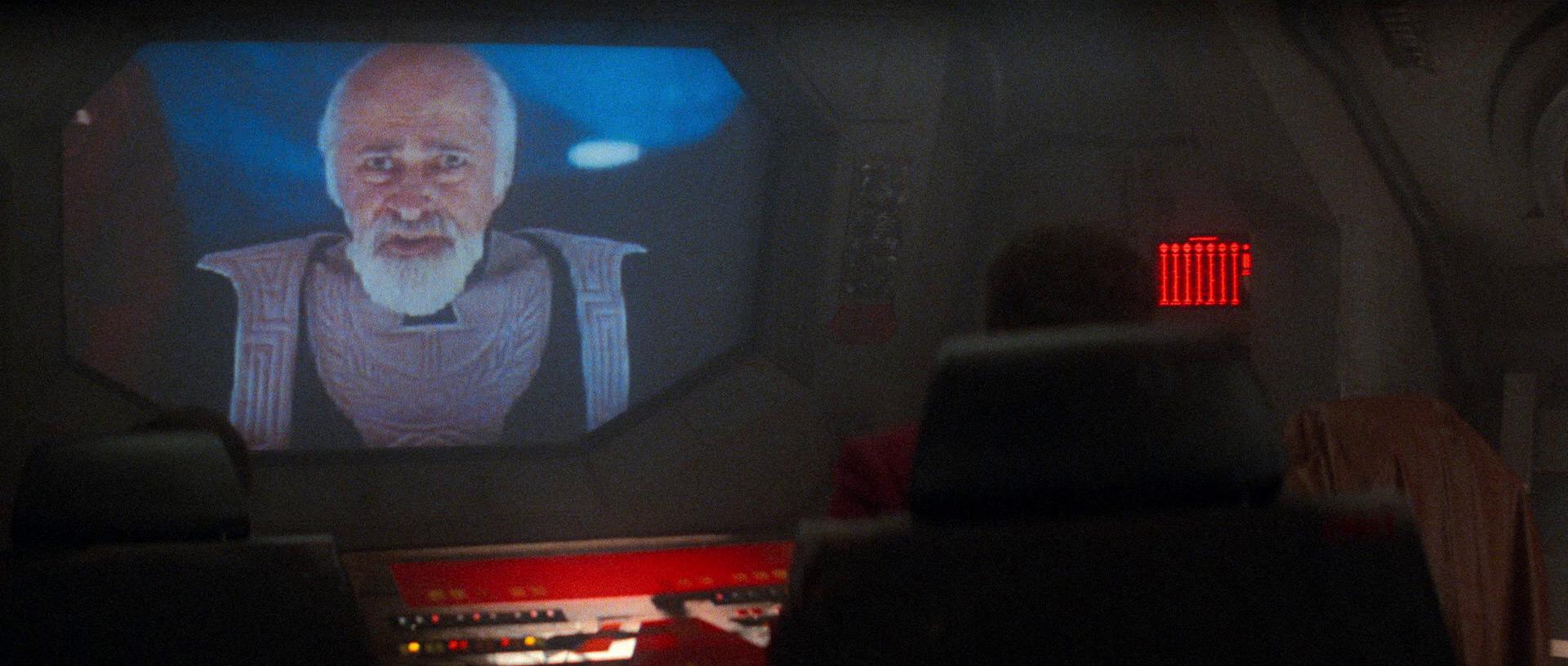 A still from the movie Star Trek IV: The Voyage Home. A large viewscreen dominates the dark bridge of a spaceship. The room is lit with red lights. On the screen, a gray haired, bearded man stares into the camera with a despondent expression on his face.