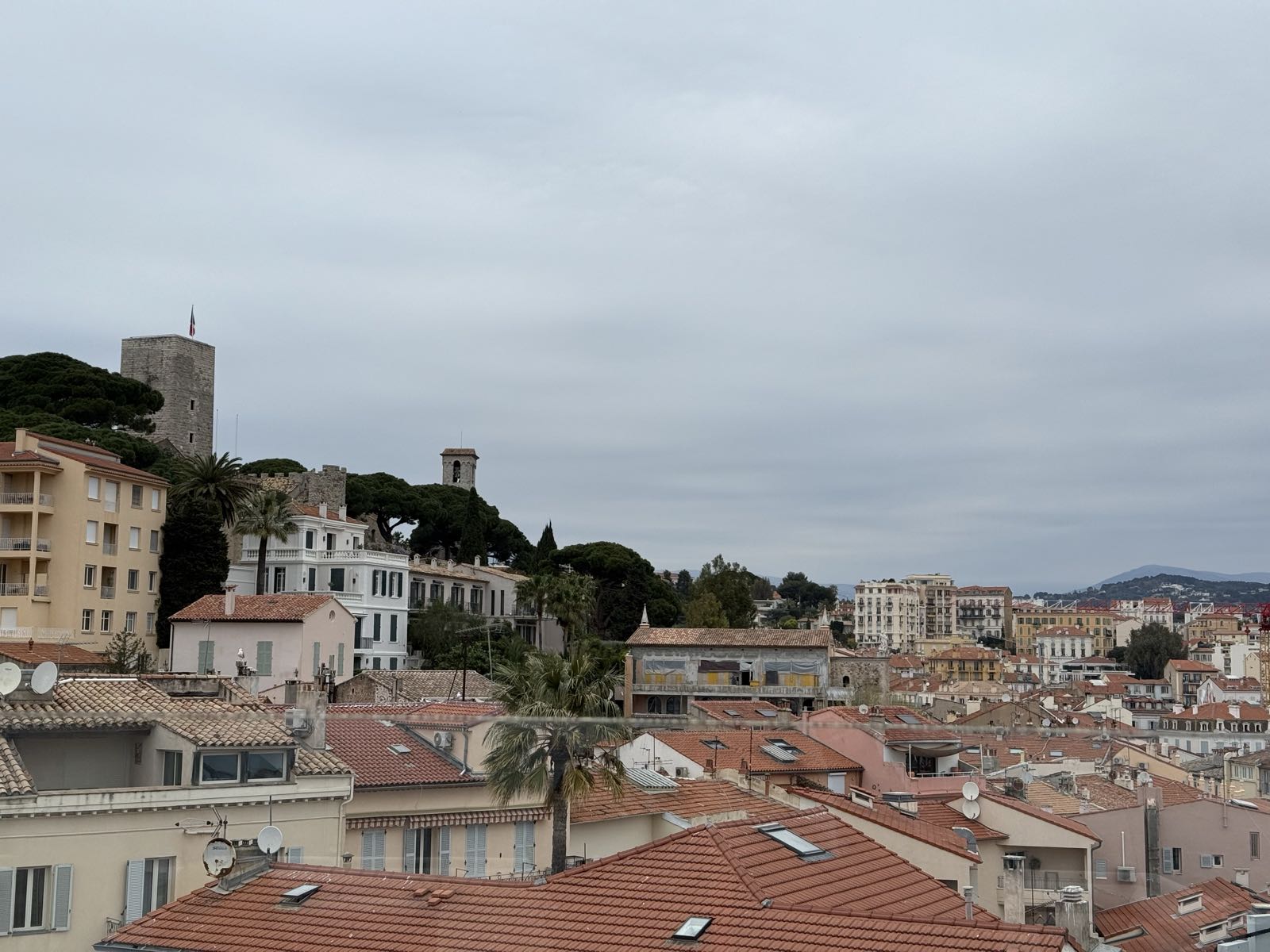 A photo of the rooftops within a Mediterranean city. Most of the roofs are made of tan/orange terracotta tiles, as is common in the region. A medieval stone tower stands on a hill in the left of the frame. The French tricolour hangs from a flagpole on the top of the tower.