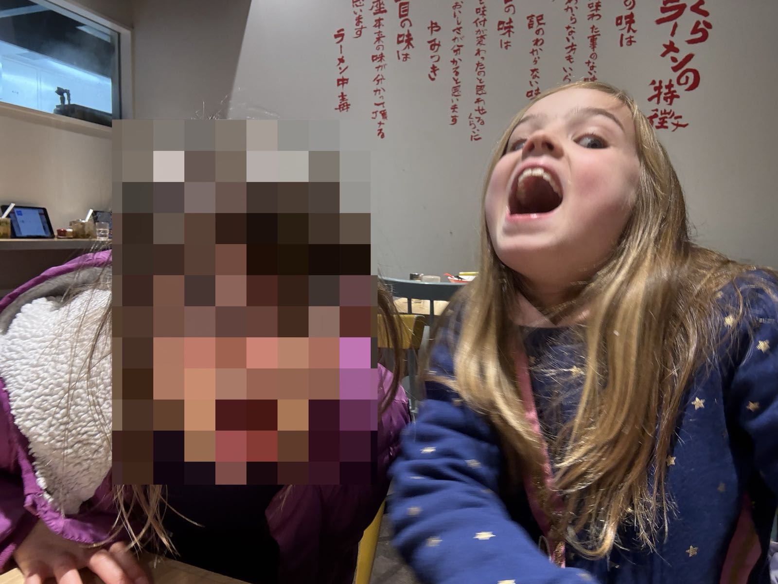 Two children are being very goofy at a table in a restaurant. Behind them, the white wall has a bunch of red text on it.