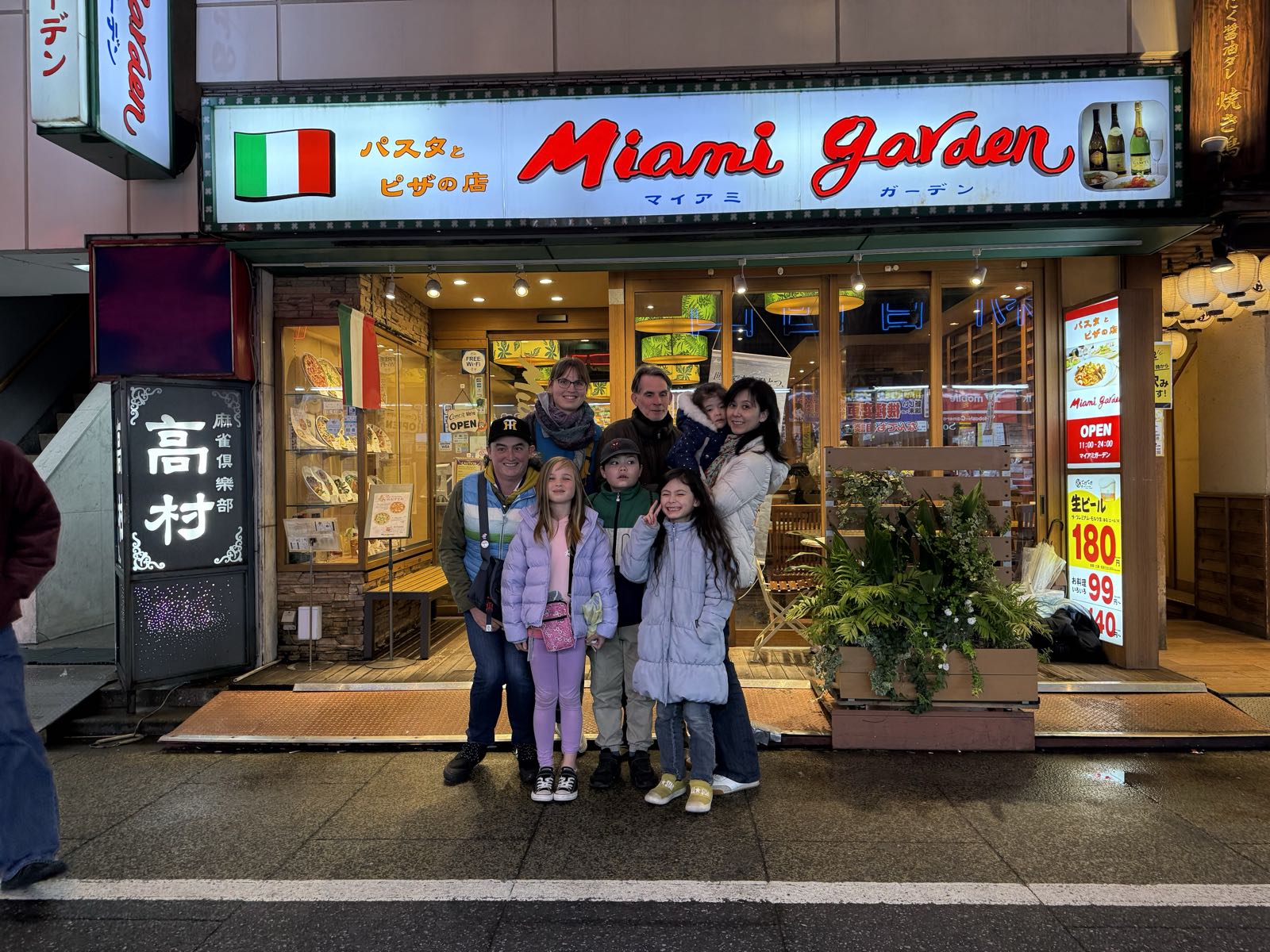 A group of adults and children pose for a photo outside of a restaurant. The restaurant has a bright sign which reads 「パスタとピザの店」 and “Miami Garden”.