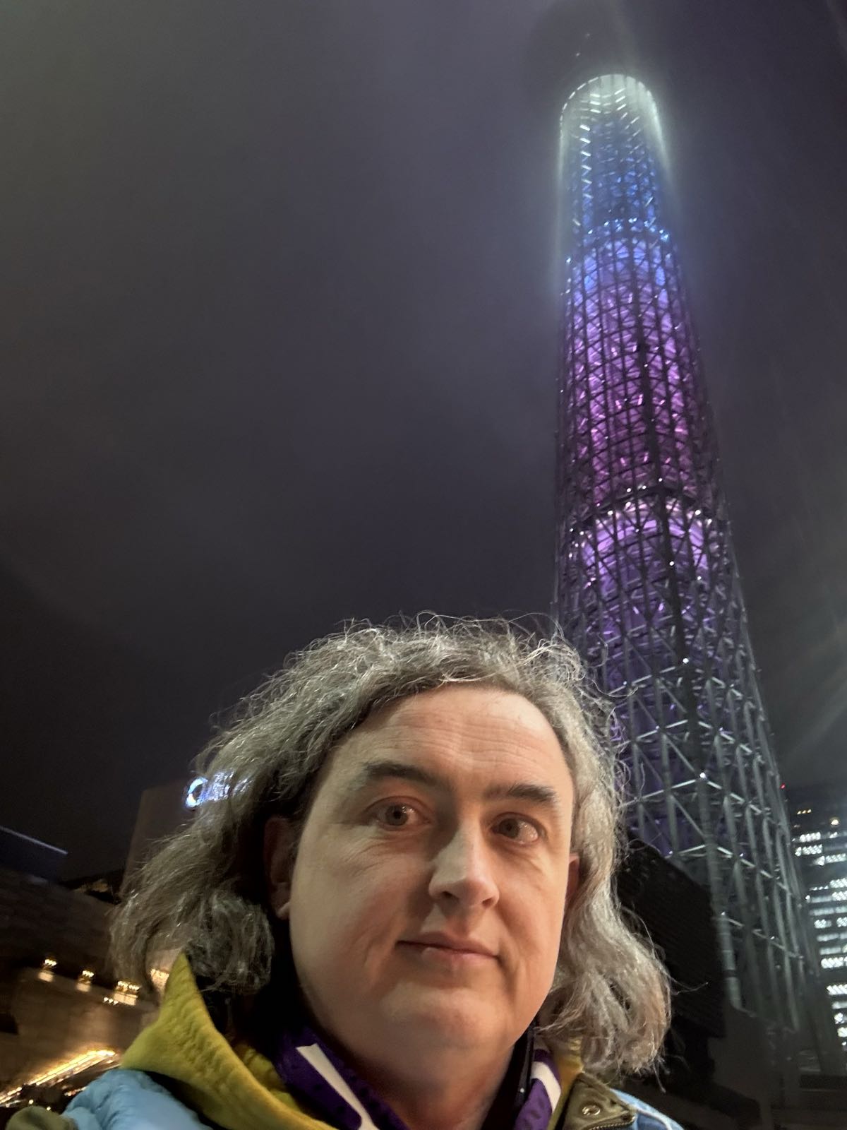 A selfie of a woman standing in front of a large, modern tower. The tower is lit up with pink, blue, and white lights. The woman has a neutral expression and shoulder-length gray hair.