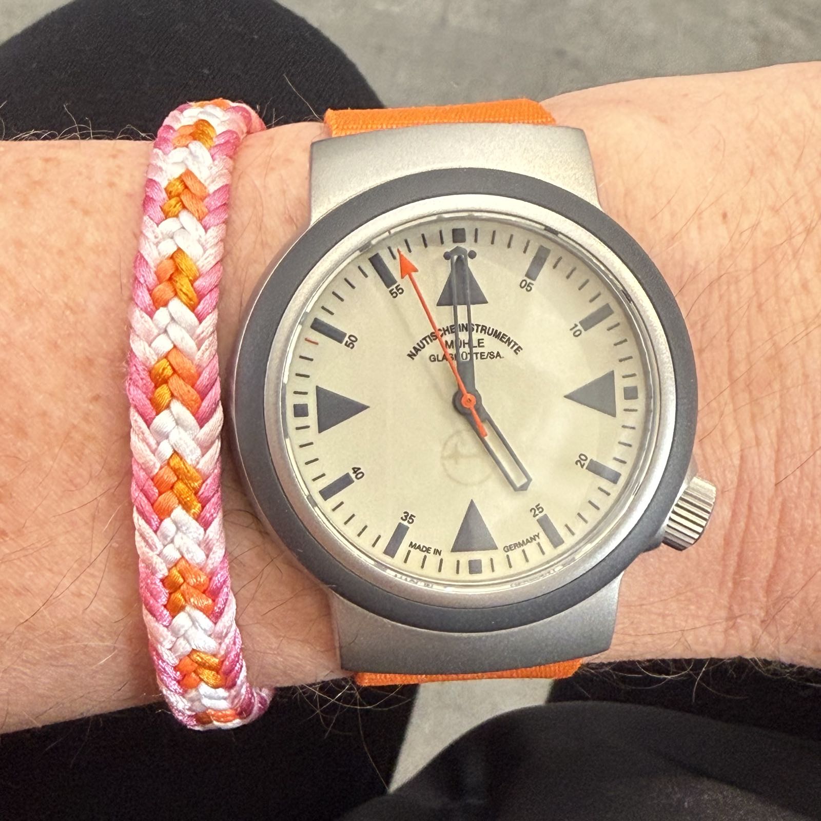 A modern-looking watch with a cream-colored dial and an orange nylon strap. The dial has triangles at 12, 3, 6, and 9 in the same gray as the bezel. The seconds hand and the tick mark at the 49 second mark are in orange.