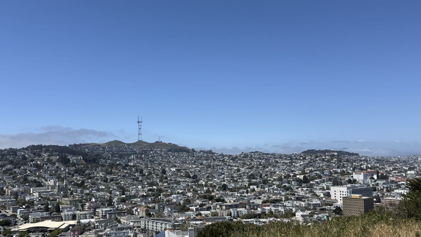 A photo of a cityscape from a hill overlooking it. Many small- and medium-sized buildings are spread uniformly across the view. The sky above the city is clear. Piercing the sky into two is a large antenna tower, which stands from a hill in the middle of town.