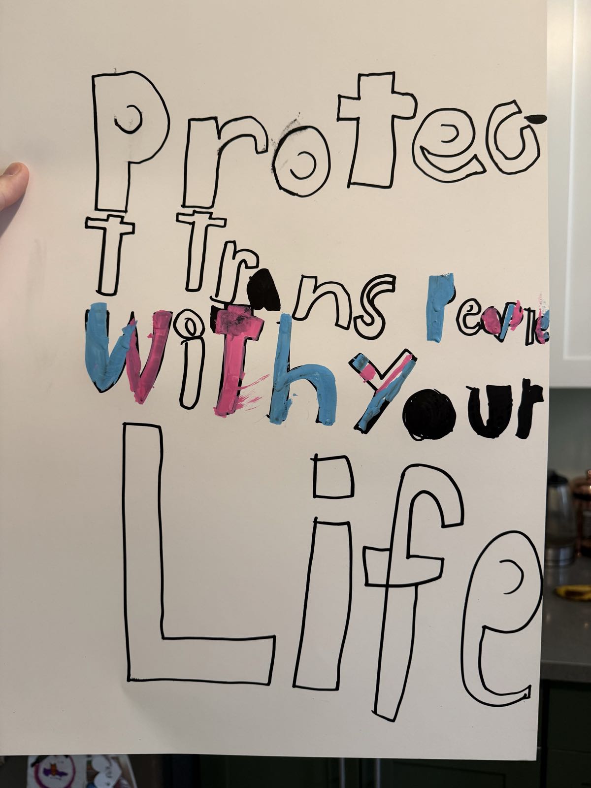 A protest sign on white posterboard reads “Protect Trans People With Your Life.” Some of the letters are colored in with blue, pink, or a combination.