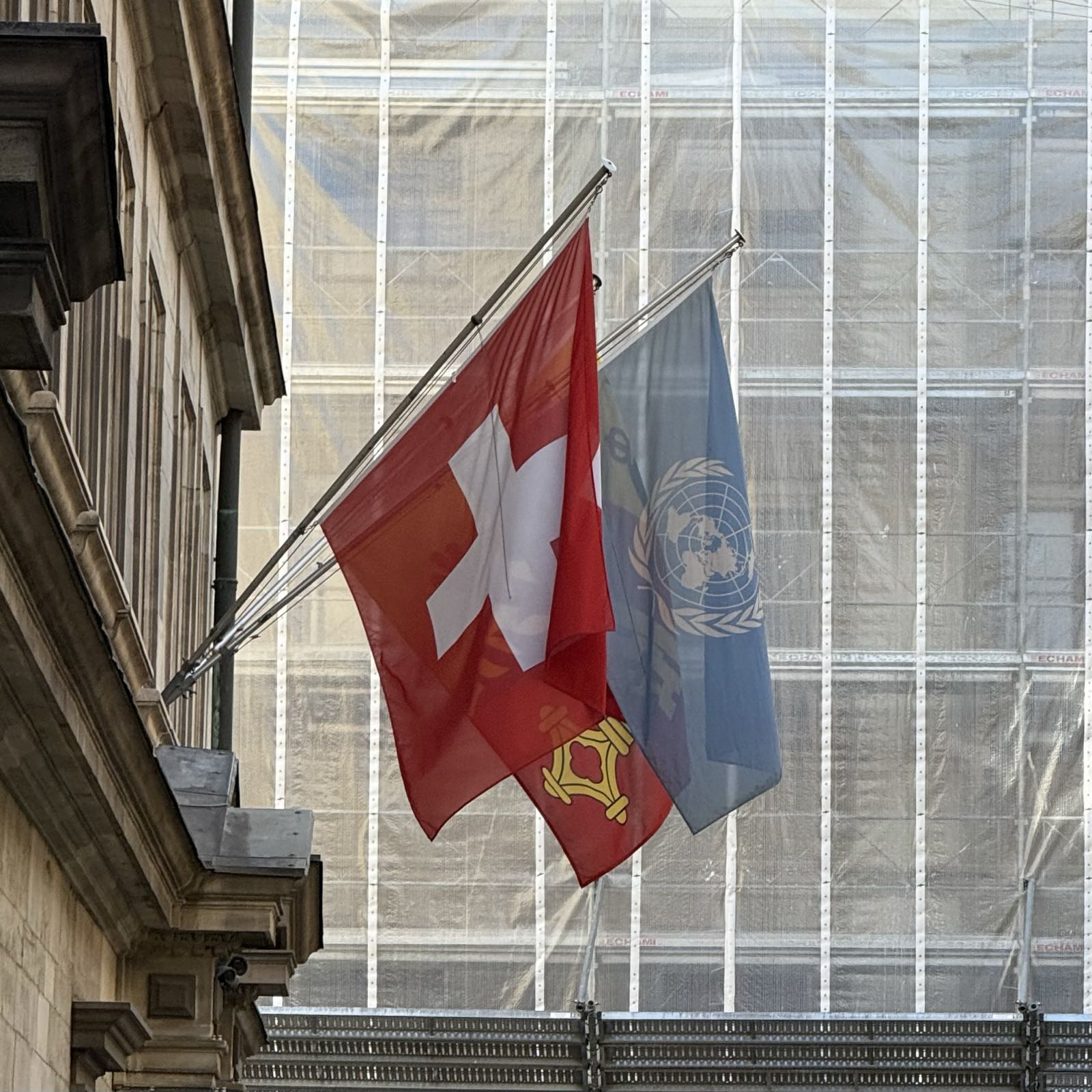Three flags hang from an old stone building: the flag of the United Nations, the flag of Switzerland, and the flag of the Canton of Geneva.