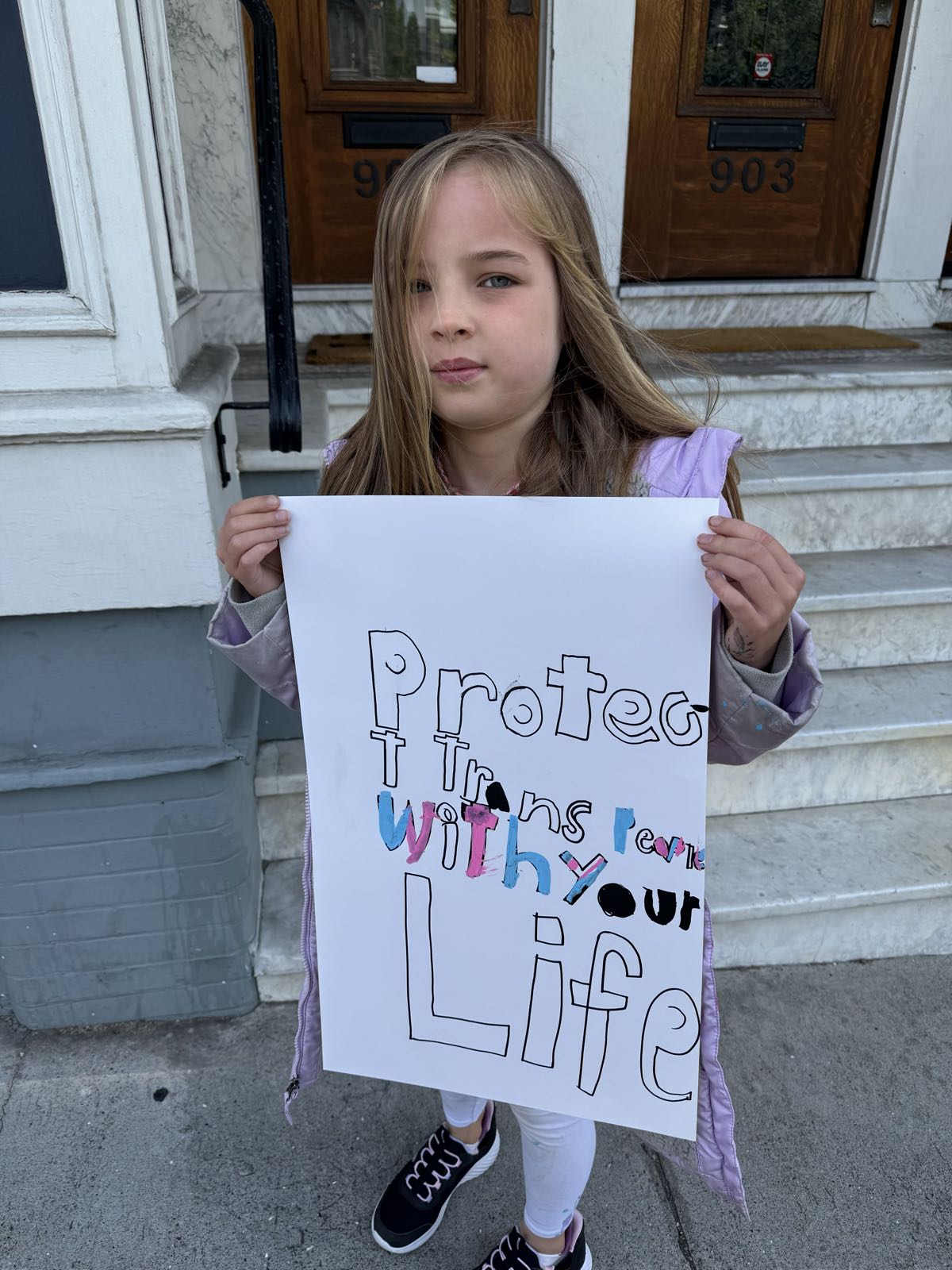 A child holds a protest sign that reads “Protect Trans People With Your Life.”