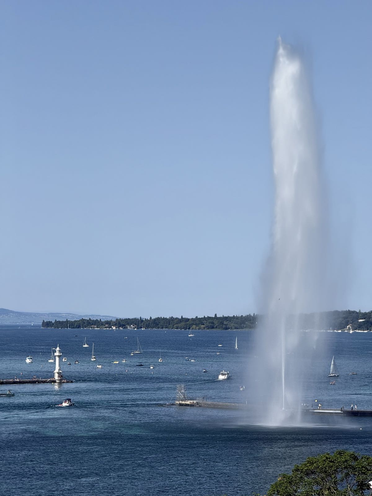 An outrageously large stream of water jets out of a lake. A wooded shoreline is visible in the distance. There are a number of boats on the water.