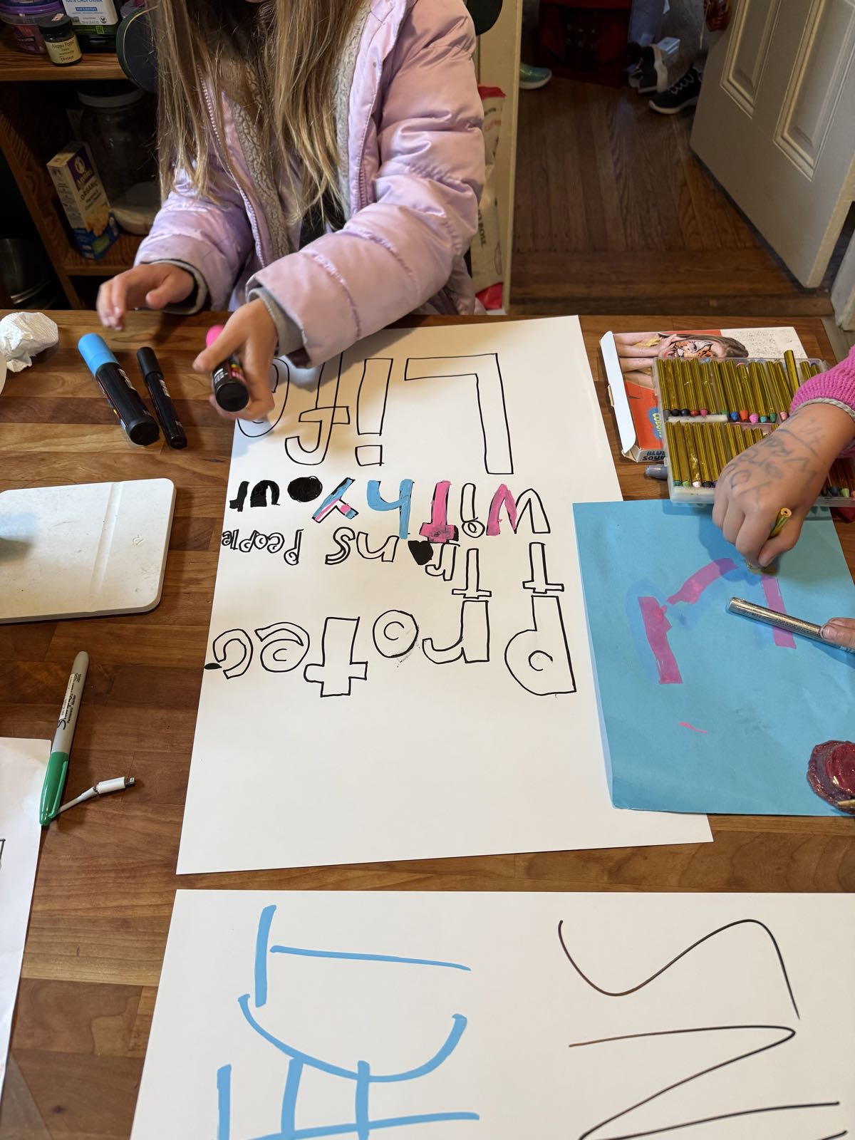 Two children sit at a table, coloring in signs with markers. One sign reads “Protect Trans People With Your Life”.