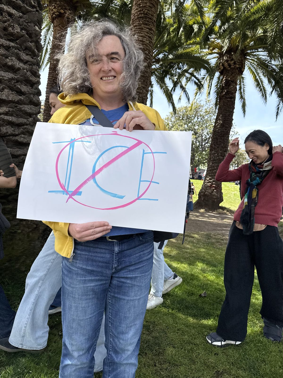 A woman in jeans and a yellow sweatshirt holds a protest sign, which reads “No ICE”.