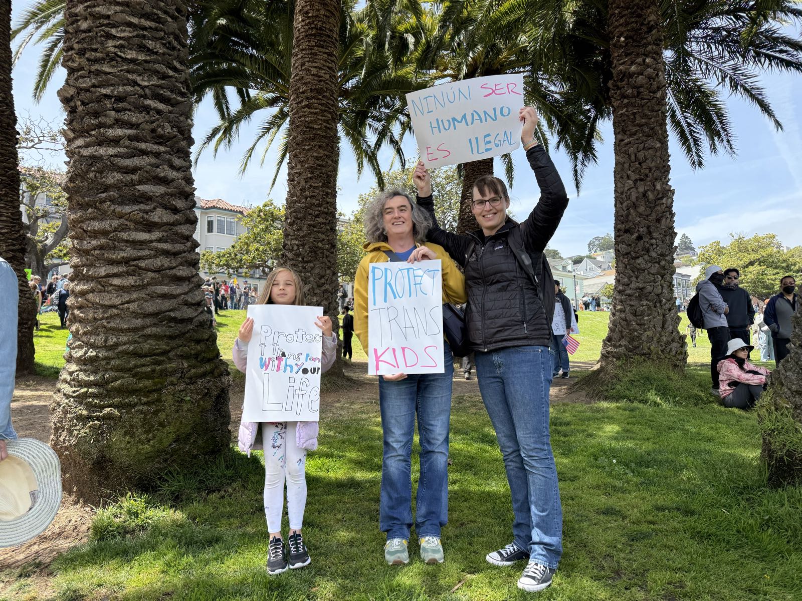 Two women and a child pose with protest signs in a park. The signs read: “Protect Trans People With Your Life”; “Protect trans kids”; and “Nin[g]ún ser humano es ilegal”.