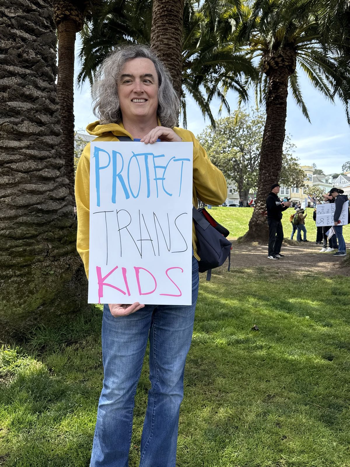 A woman in jeans and a yellow sweatshirt holds a protest sign, which reads “Protect trans kids”.
