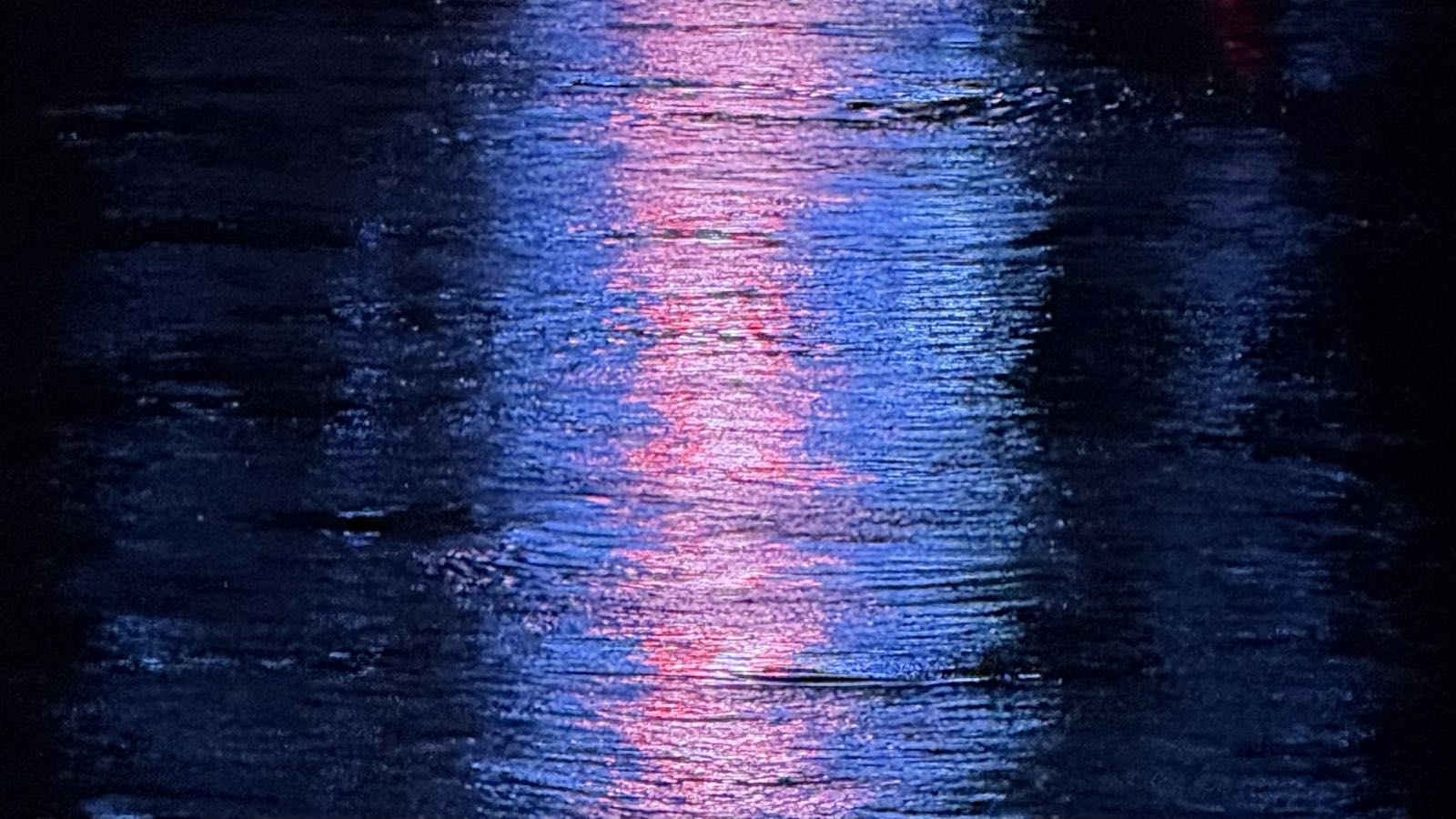 Blue, pink, and white lights reflect off of a body of water at night, inadvertently recreating the transgender pride flag.