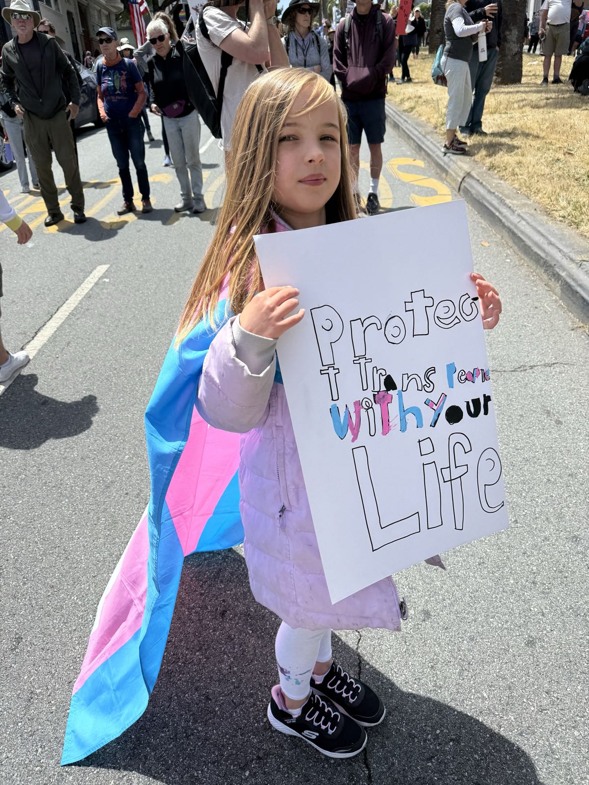 A child holds a protest sign that reads “Protect Trans People With Your Life.” She’s wearing the transgender pride flag as a cape.