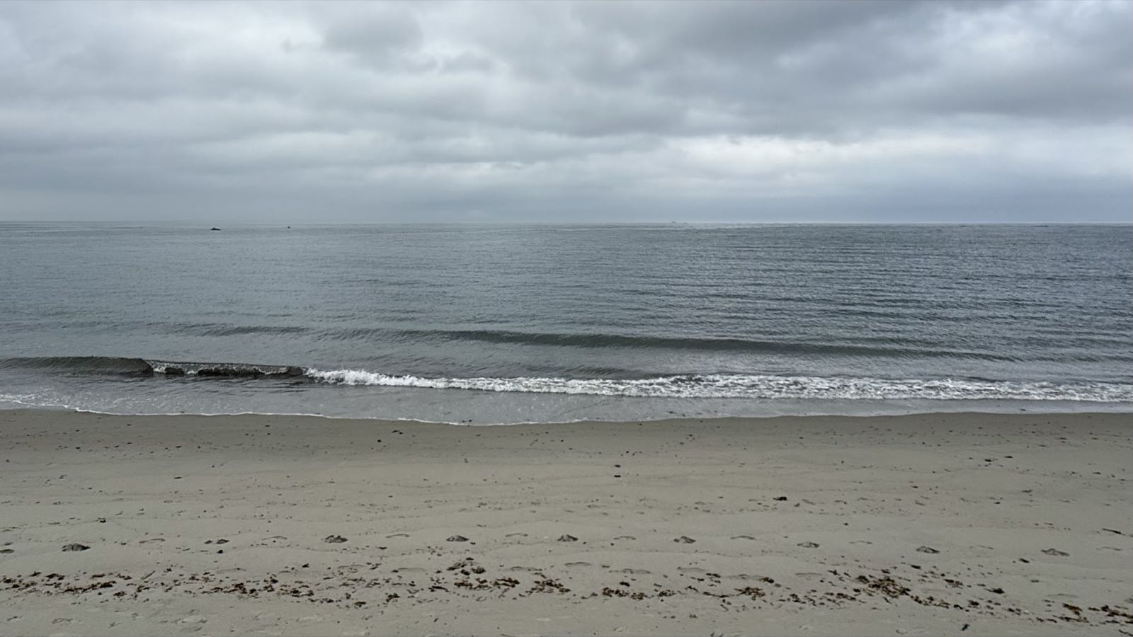 Small waves from a calm ocean break on a beach under a cloudy sky.