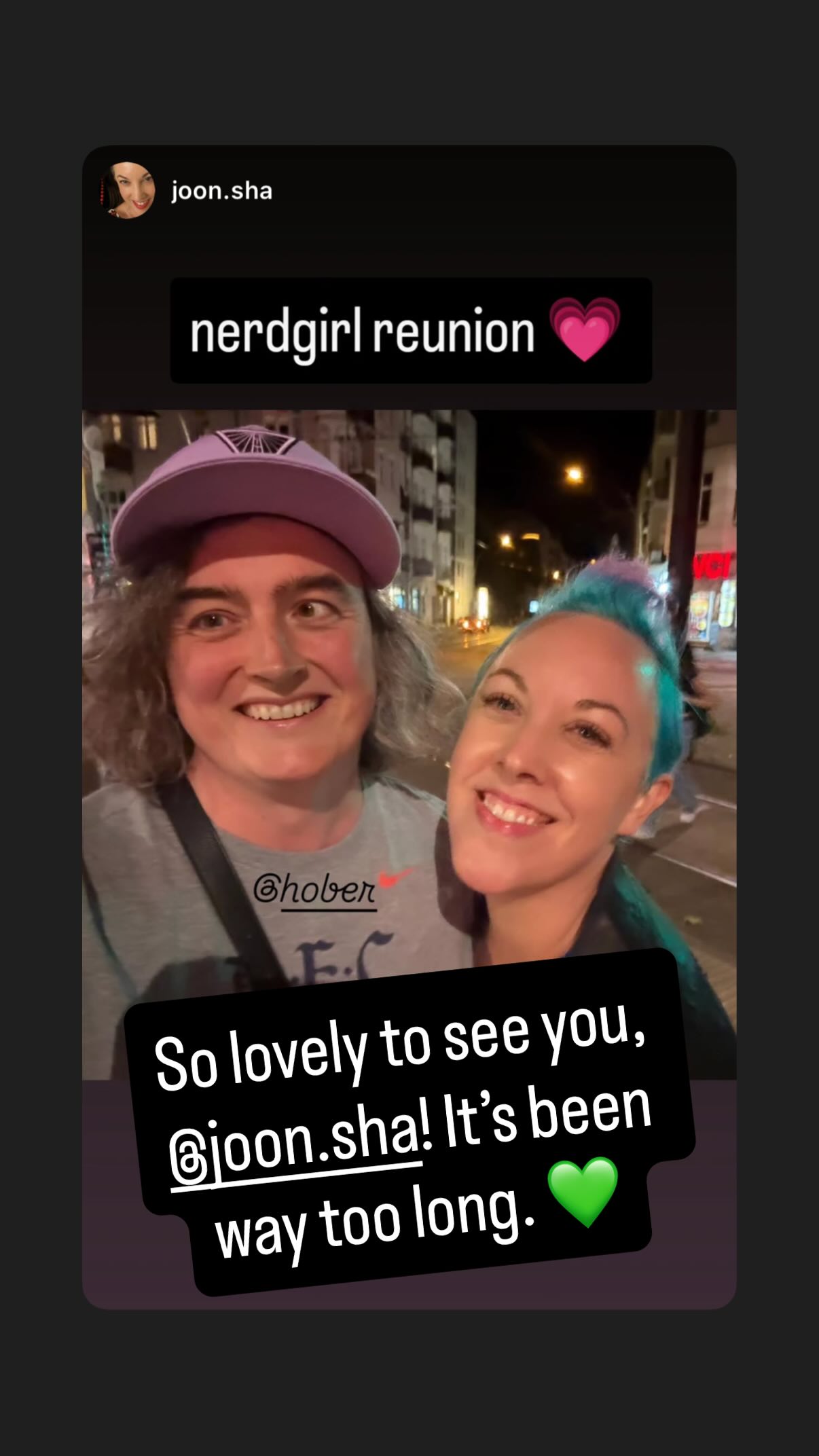 A woman in a lavender hat poses for a selfie with a woman with blue hair. They both seem very happy to see each other.