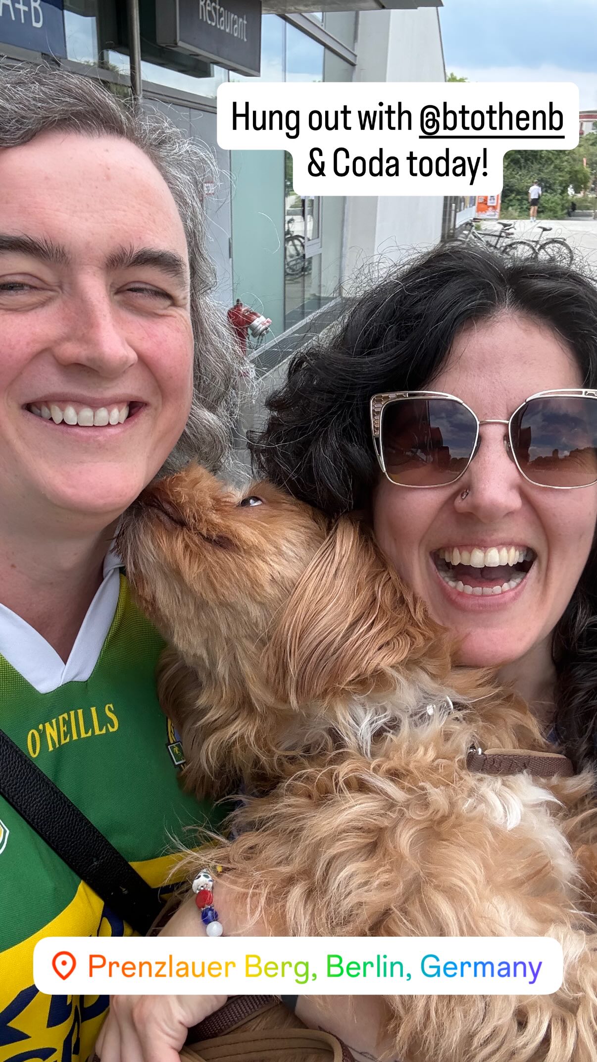 Two smiling women pose for a selfie. The woman on the right is holding a brown dog, who is licking the face of the woman on the left.