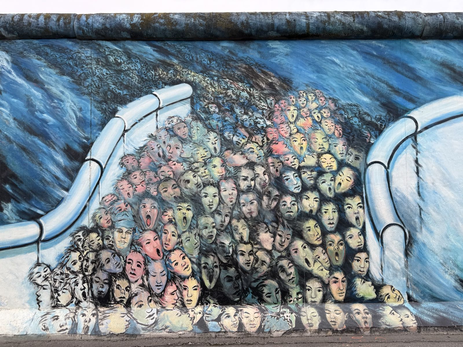 A teeming mass of faces flow through a break in a wall, as if the faces are part of a river of humanity.