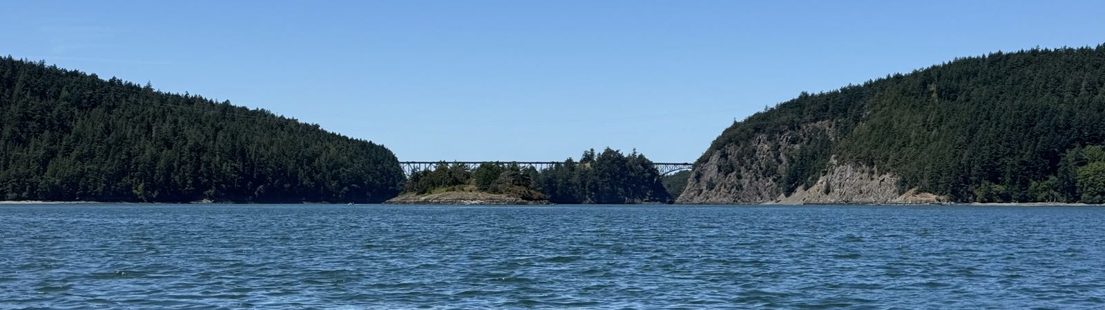 Two large, wooded land masses are connected with a bridge. The bridge appears to pass over a third, smaller land mass, but it’s a little hard to tell from the angle of view. There’s ocean between the camera and the bridge, and a clear sky above.