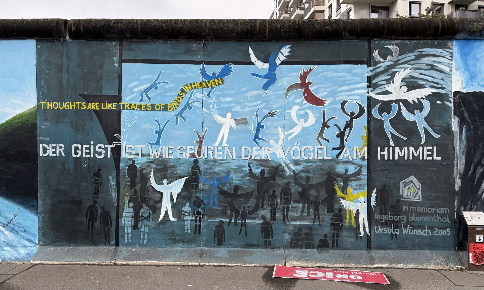 A painting depicts a dark scene below a bright blue sky. People, some with angel wings, are in various states of distress, or are flying away. A message appears in both German and English: „Der Geist ist wie Spuren der Vögel am Himmel,“ and “Thoughts are like traces of birds in Heaven.”
