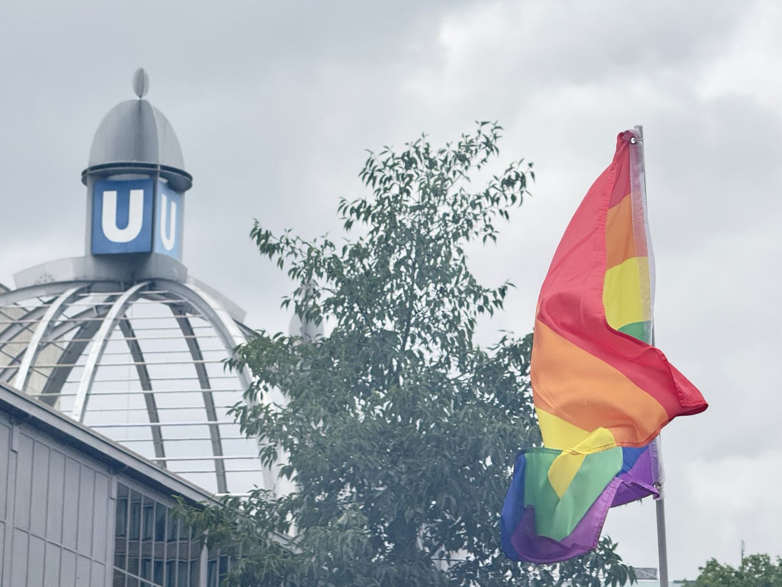 On the left, a dome is topped with a large letter “U”. On the right, a large Pride flag flies.