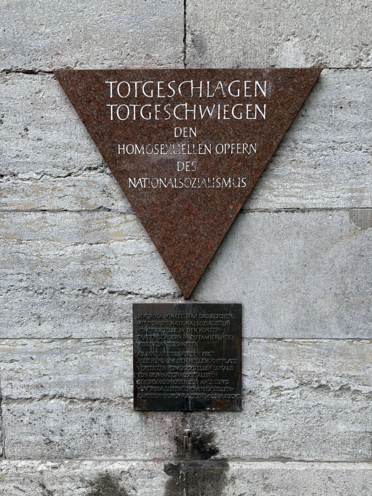 A plaque on a concrete wall is triangle-shaped, and reads “TOTGESCHLAGEN TOTGESCHWIEGEN DEN HOMOSEXUELLEN OPFERN DES NATIONALSOZIALISMUS” in German.
