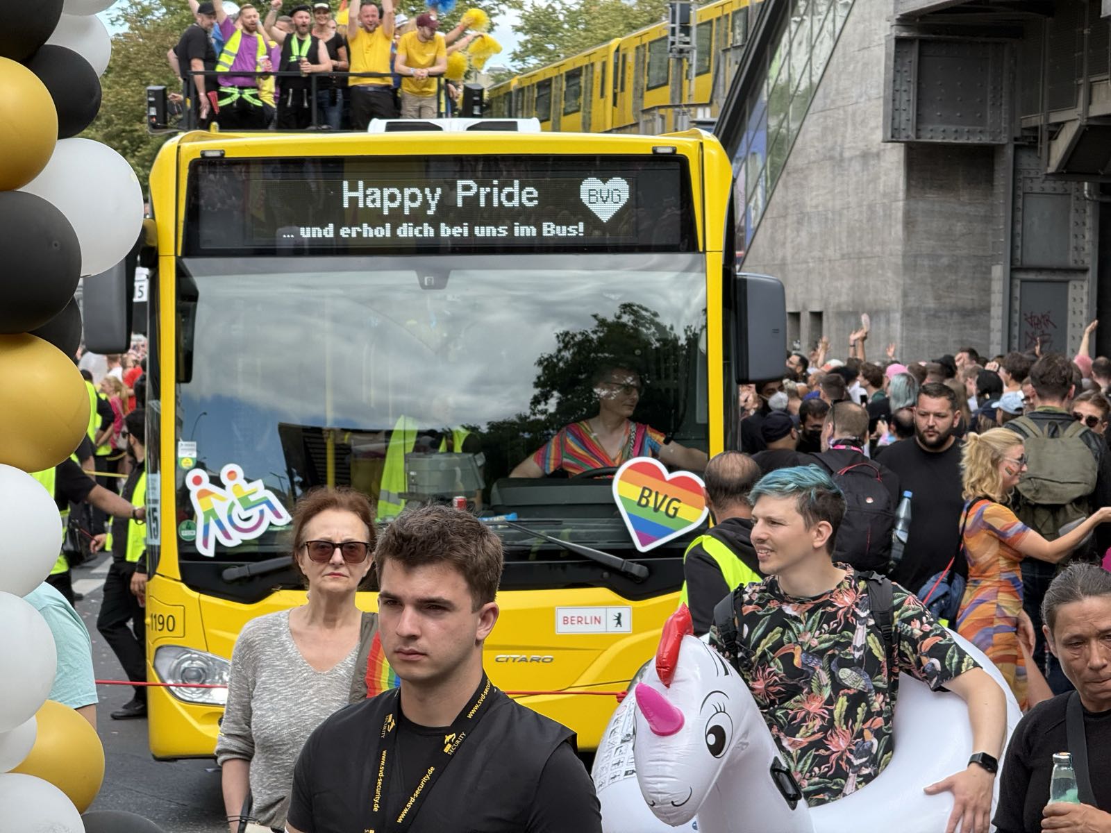 A yellow bus drives toward the camera, among a large crowd. The bus’ overhead sign reads “Happy Pride und erhol dich bei uns im Bus!”
