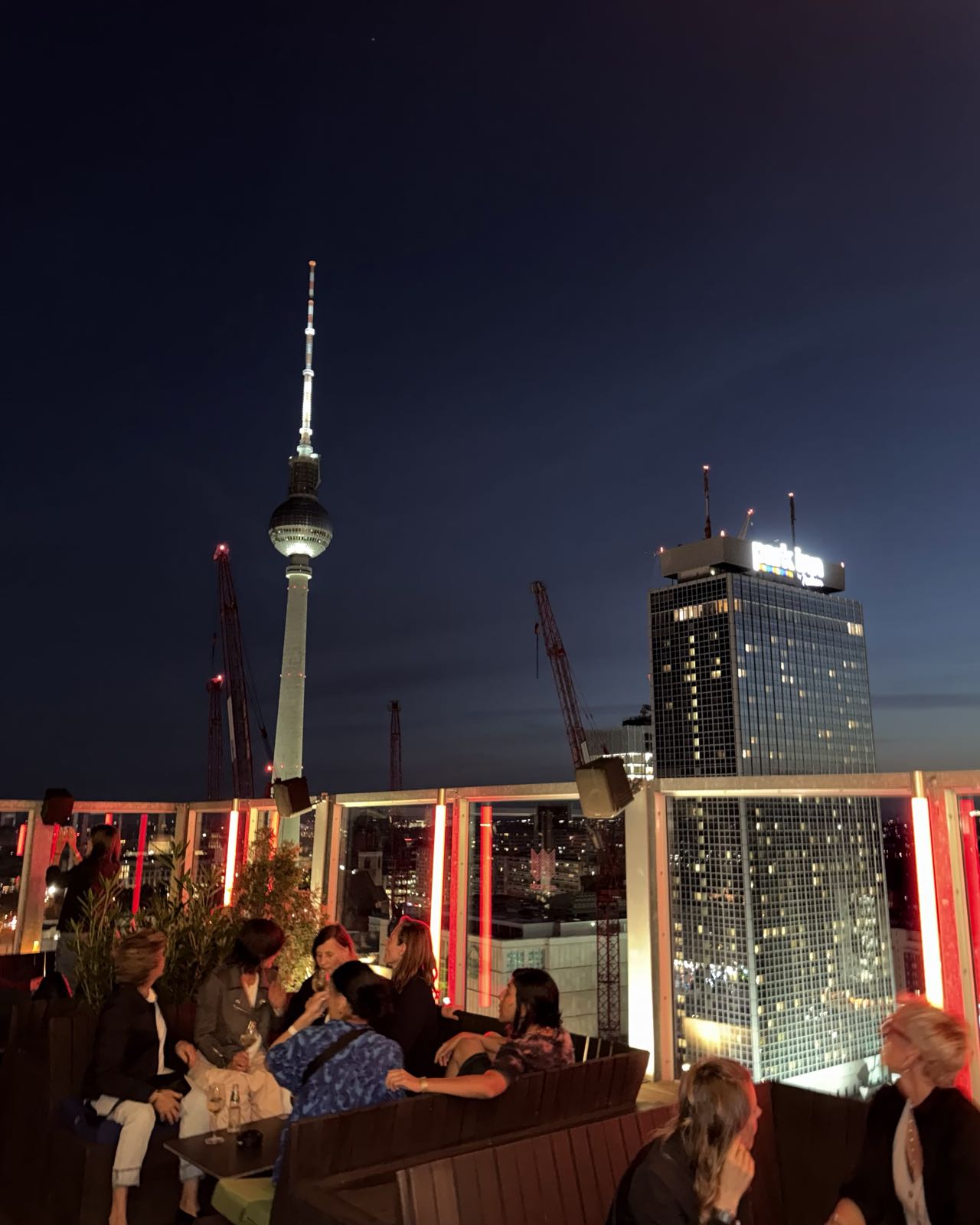 A bunch of women are sitting around tables on a rooftop at night. Behind them, Berlin’s Fernsehturm is visible.
